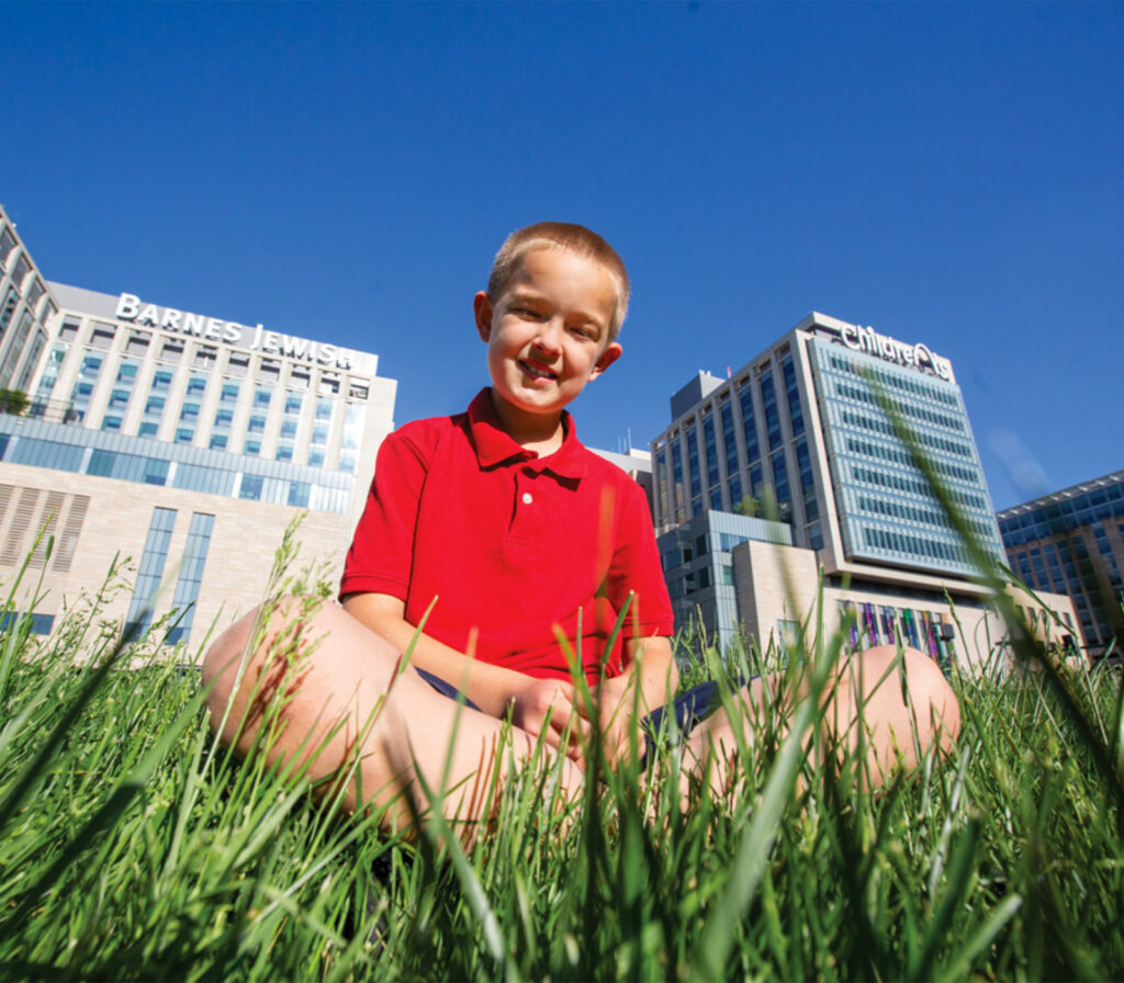 Photo of Noah Hingst sitting in grass in front of Barnes-Jewish Hospital and St. Louis Children’s Hospital.